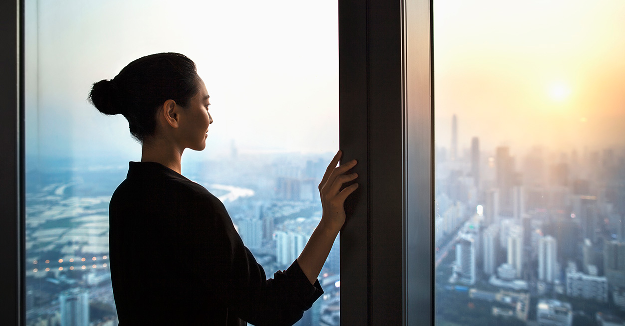 Una donna d’affari che guarda fuori da una grande finestra verso un paesaggio urbano durante il tramonto